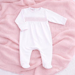 A white long-sleeve baby footed romper with a pink smocked chest band, laid flat on a textured pink muslin blanket.