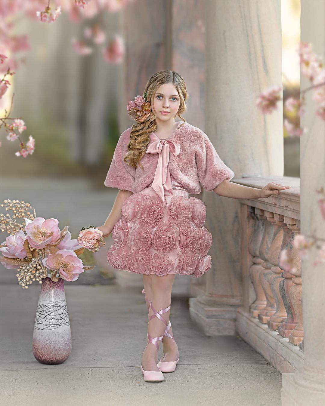 girl in a pink fluffy bolero jacket faux fur with floral skirt standing in a decorative setting with flowers and columns.